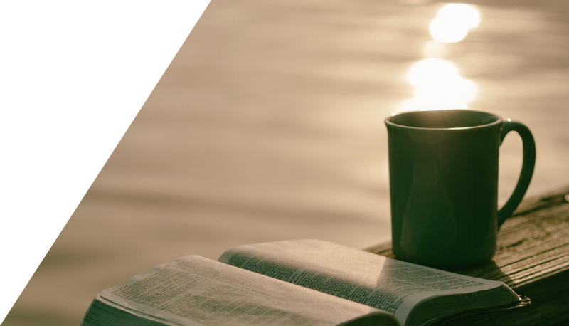 A cup of coffee and a Bible on a wooden railing by a sunny lake